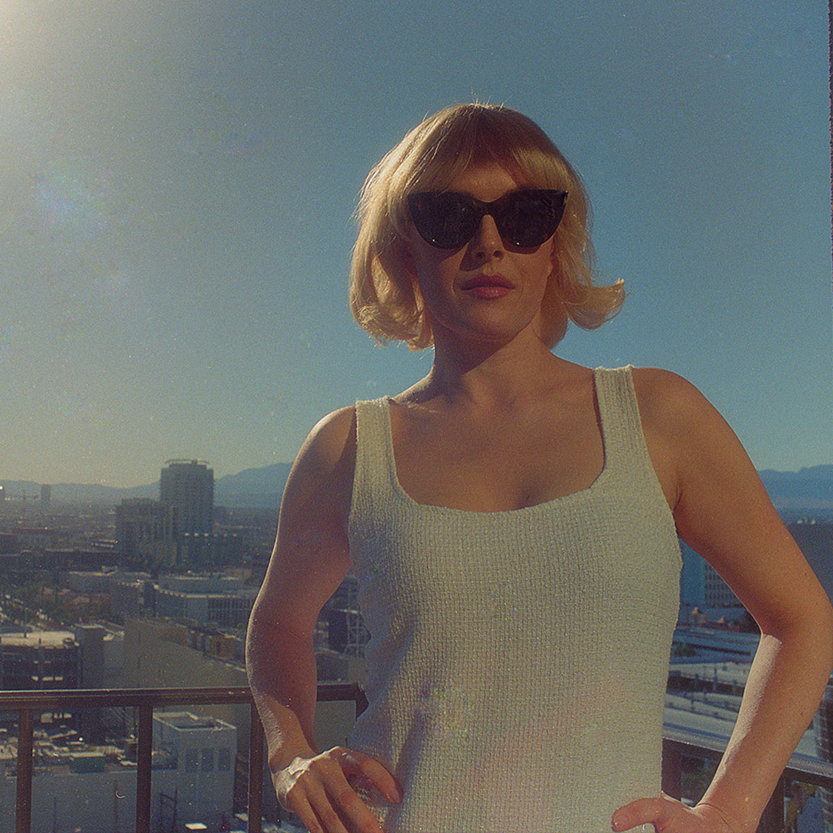 Gwenno stands high up in front of a city scape in a white dress and sunglasses