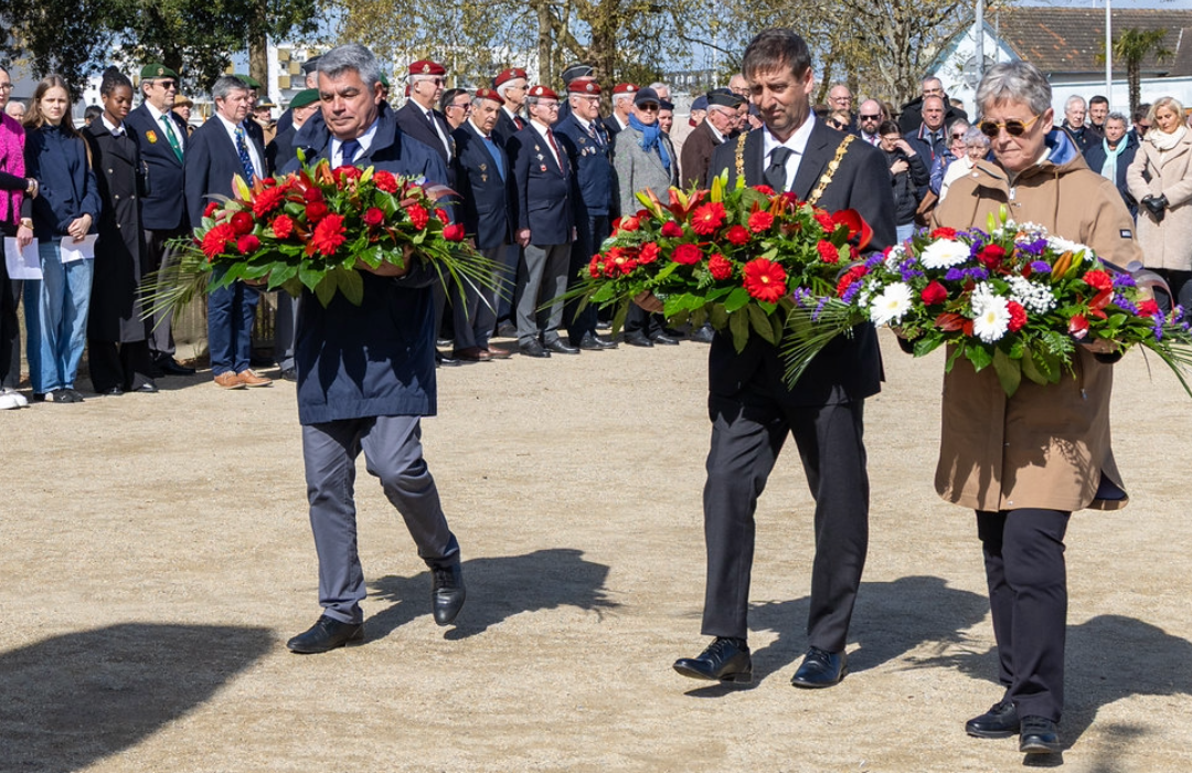 The Mayor of Falmouth, Cllr Alan Jewell, alongside two French officials, carry floral wreaths towards a memorial during a remembrance ceremony in Saint-Nazaire, Brittany, marking 84 years since Operation Chariot, a World War II Allied raid in 1942, while attendees and veterans stand behind them.